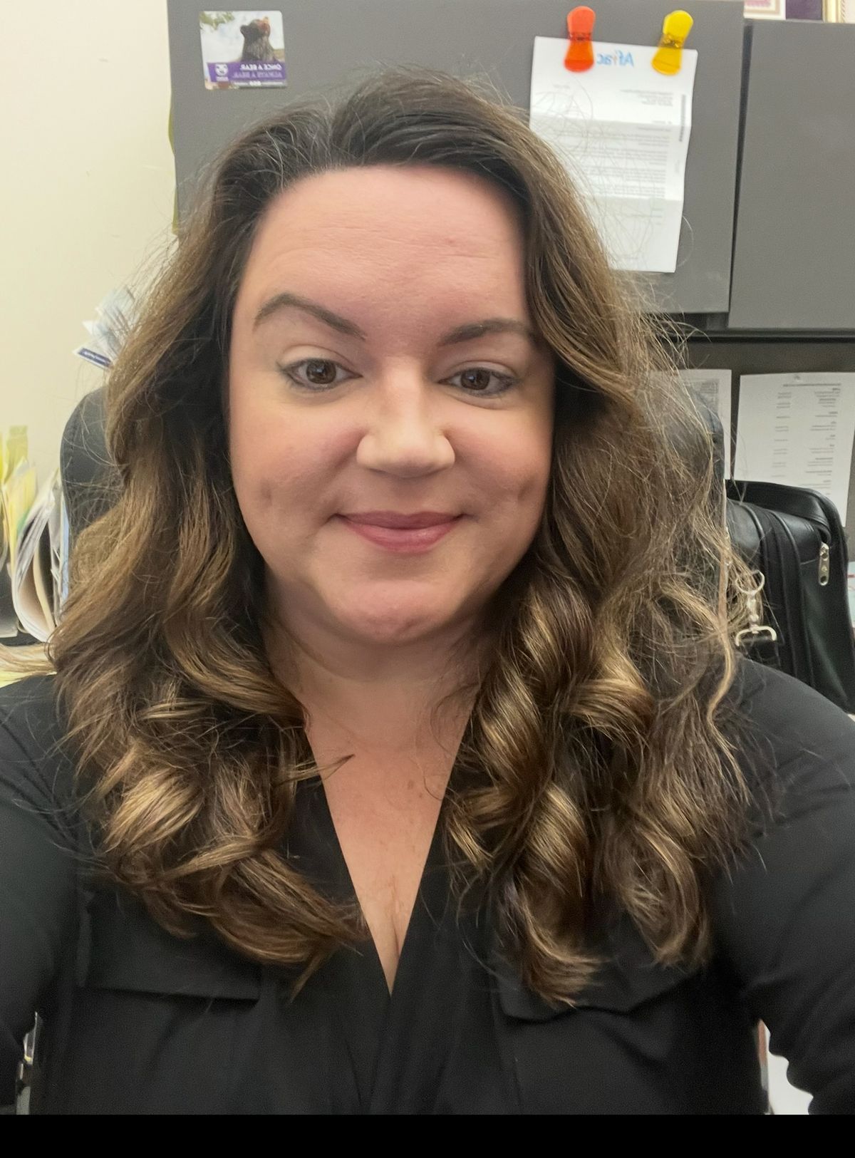 A woman with long, wavy brown hair, wearing a black shirt and smiling in an office setting.