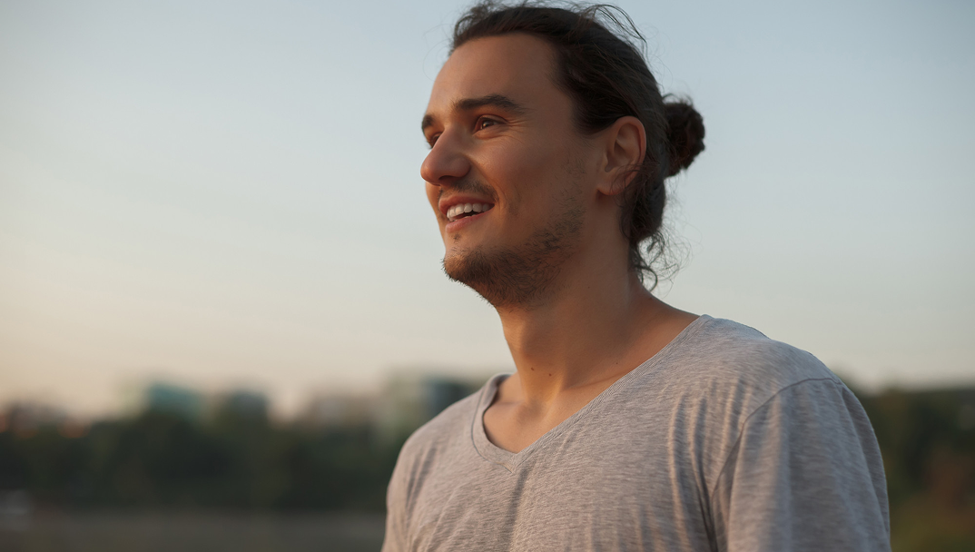 Smiling person with long hair in a bun, wearing a gray shirt outdoors.