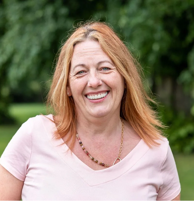 Woman smiling outdoors with greenery in the background.