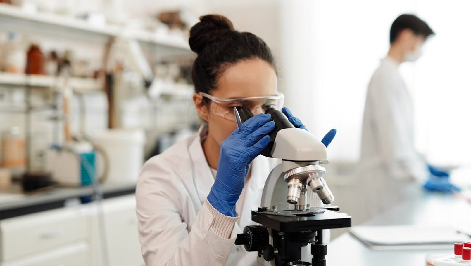 Person using a microscope in a laboratory, wearing gloves and goggles.