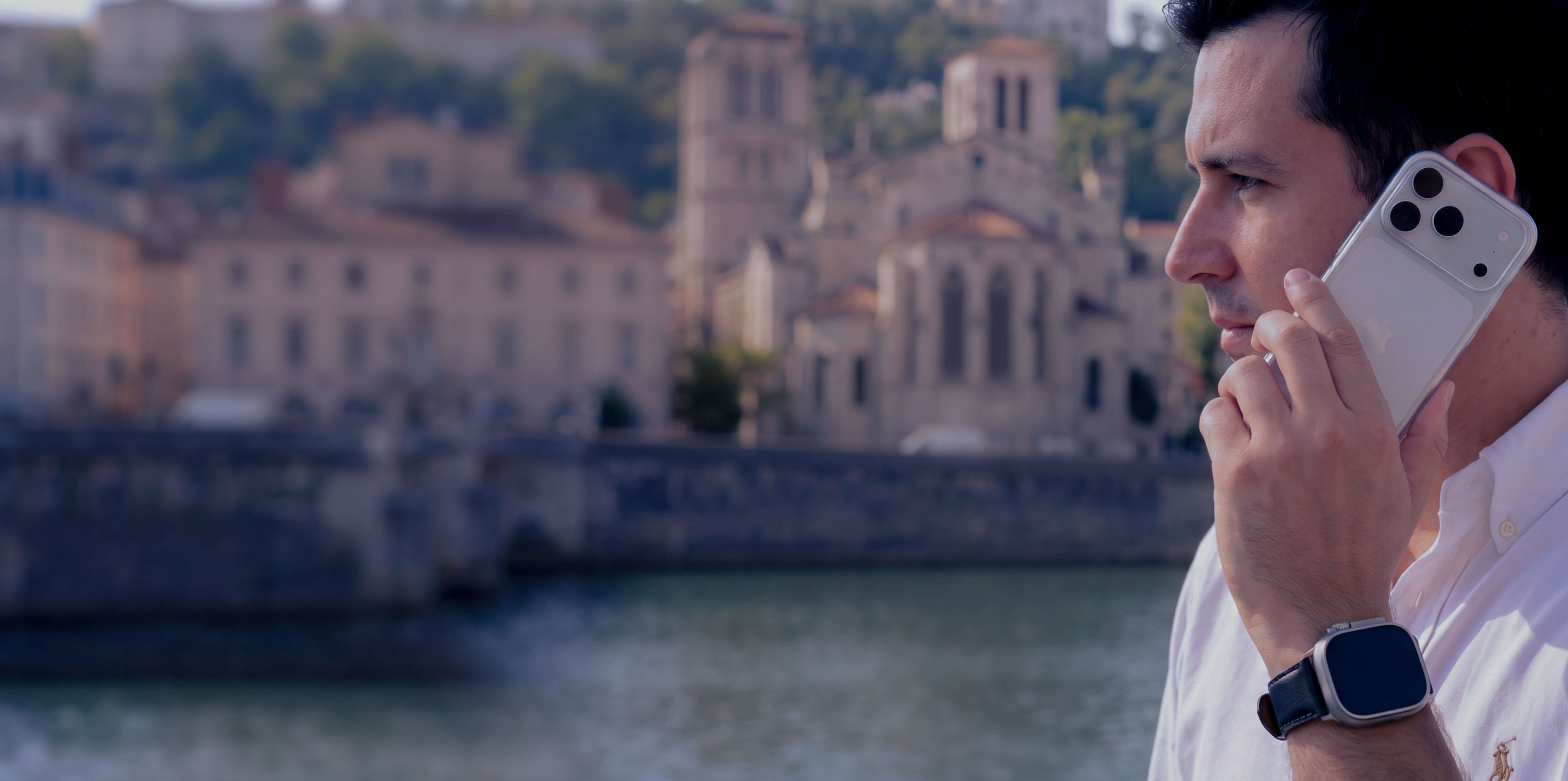 A man in profile holding a smartphone to his ear, with a blurry European city in the background.