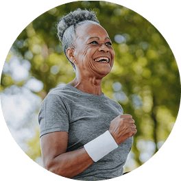 An older Black woman with gray hair smiles while jogging outdoors in a park.