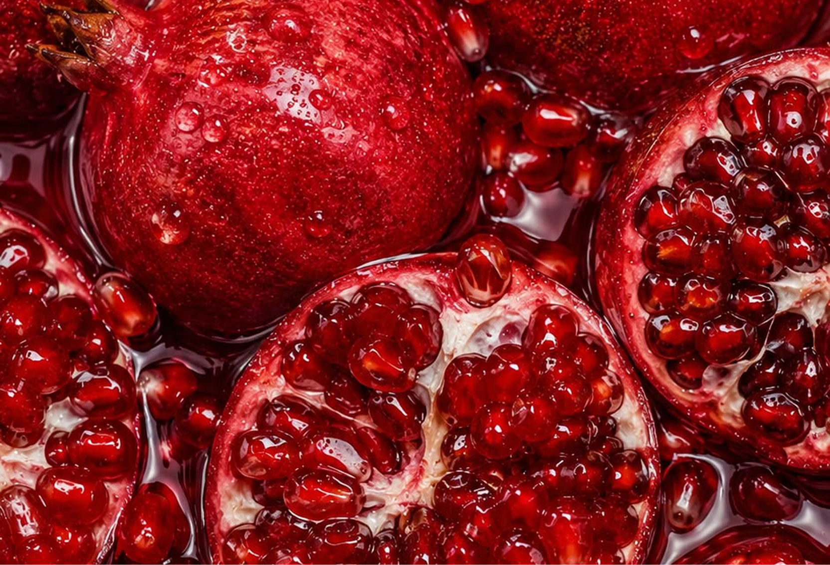 A close-up of whole and halved pomegranates, wet with juice and showing their glistening red seeds.