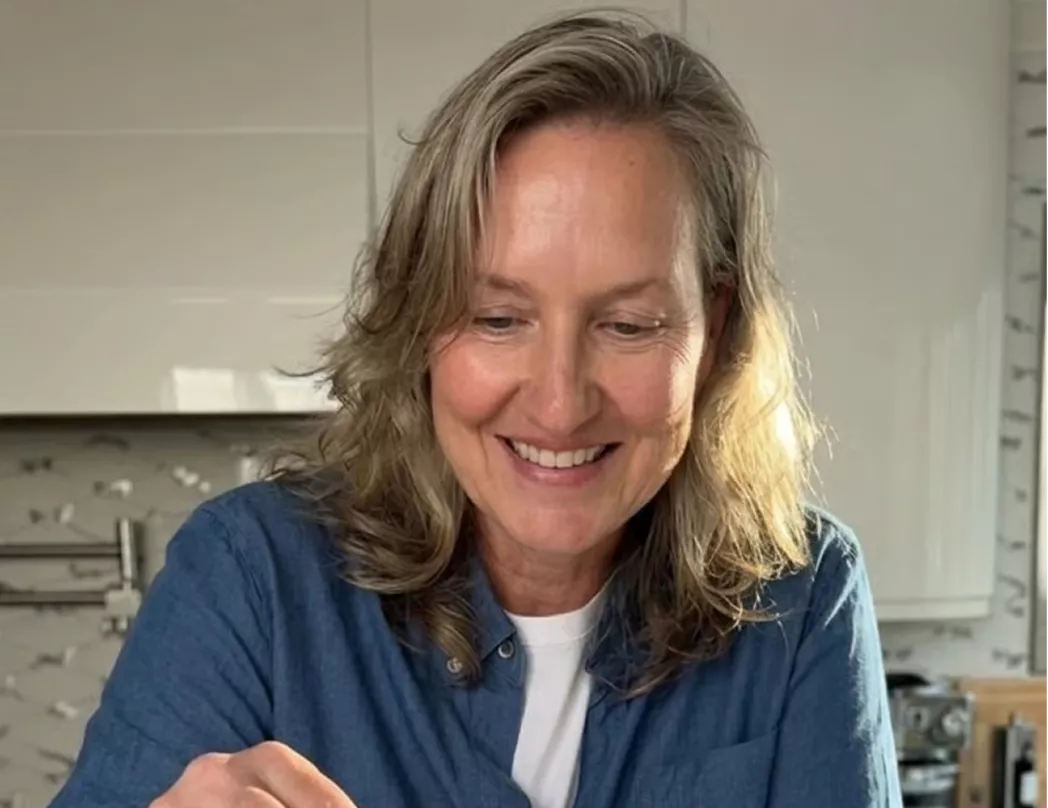 A smiling woman with wavy, blonde hair wearing a blue shirt over a white t-shirt in a kitchen.
