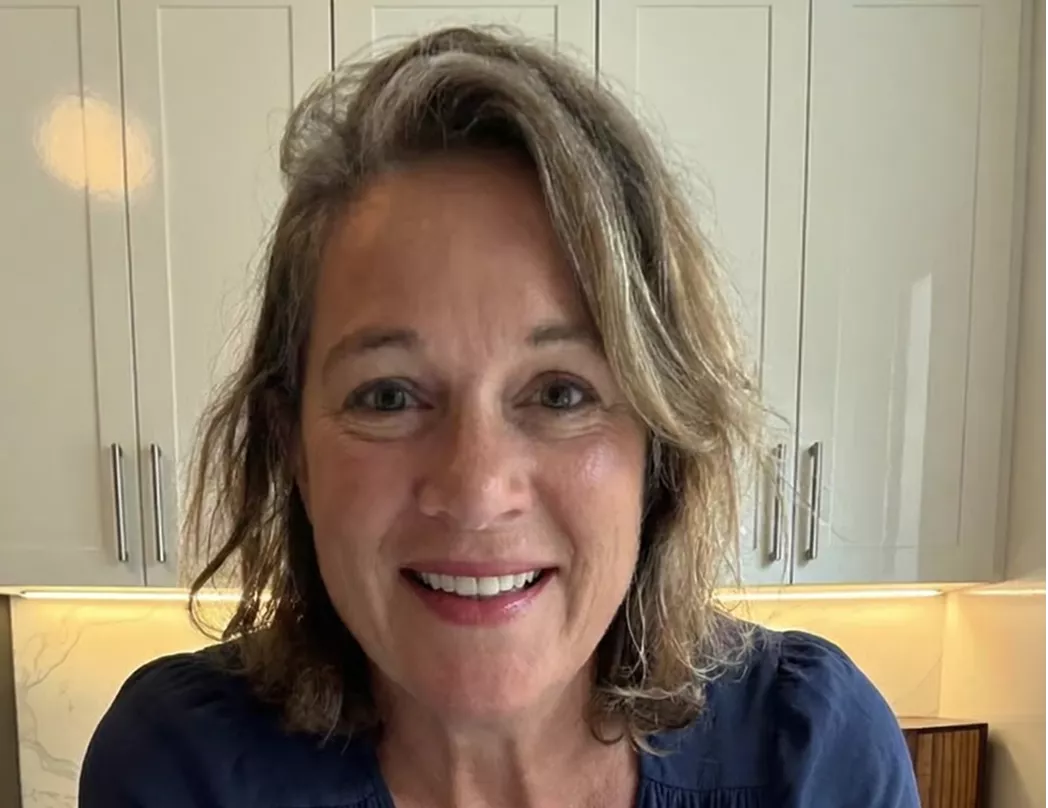 A smiling, middle-aged white woman with wavy, shoulder-length brown hair, wearing a blue top in a kitchen.