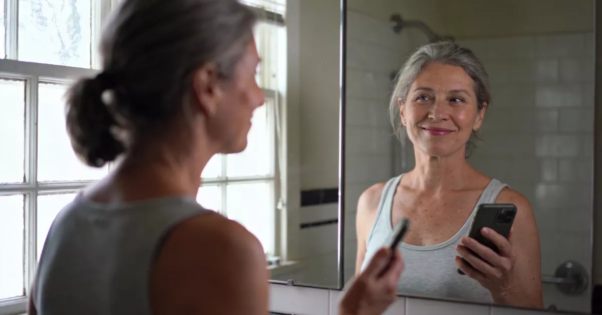 A woman with gray hair smiles at her reflection in a bathroom mirror while holding a phone.