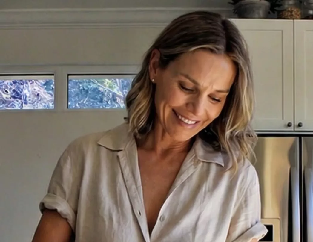 A smiling woman with blonde, shoulder-length hair looks down while wearing a beige shirt in a kitchen.