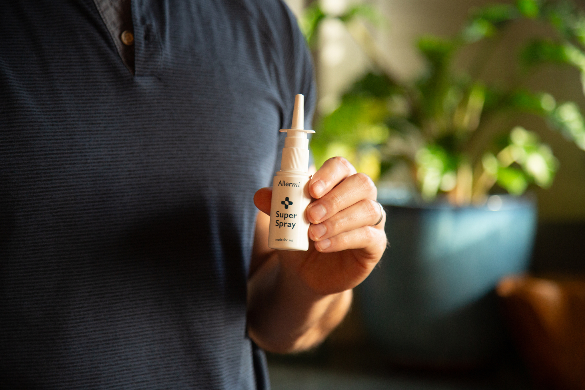 A person in a striped shirt holds up a small, white bottle of Allermi Super Spray.