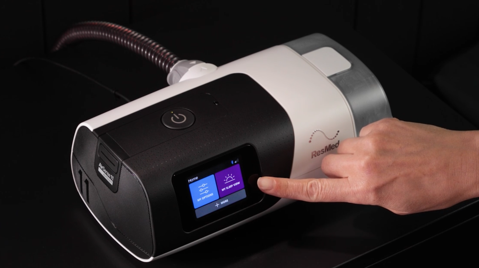 A hand operates the touchscreen on a white and black ResMed CPAP machine on a dark surface.