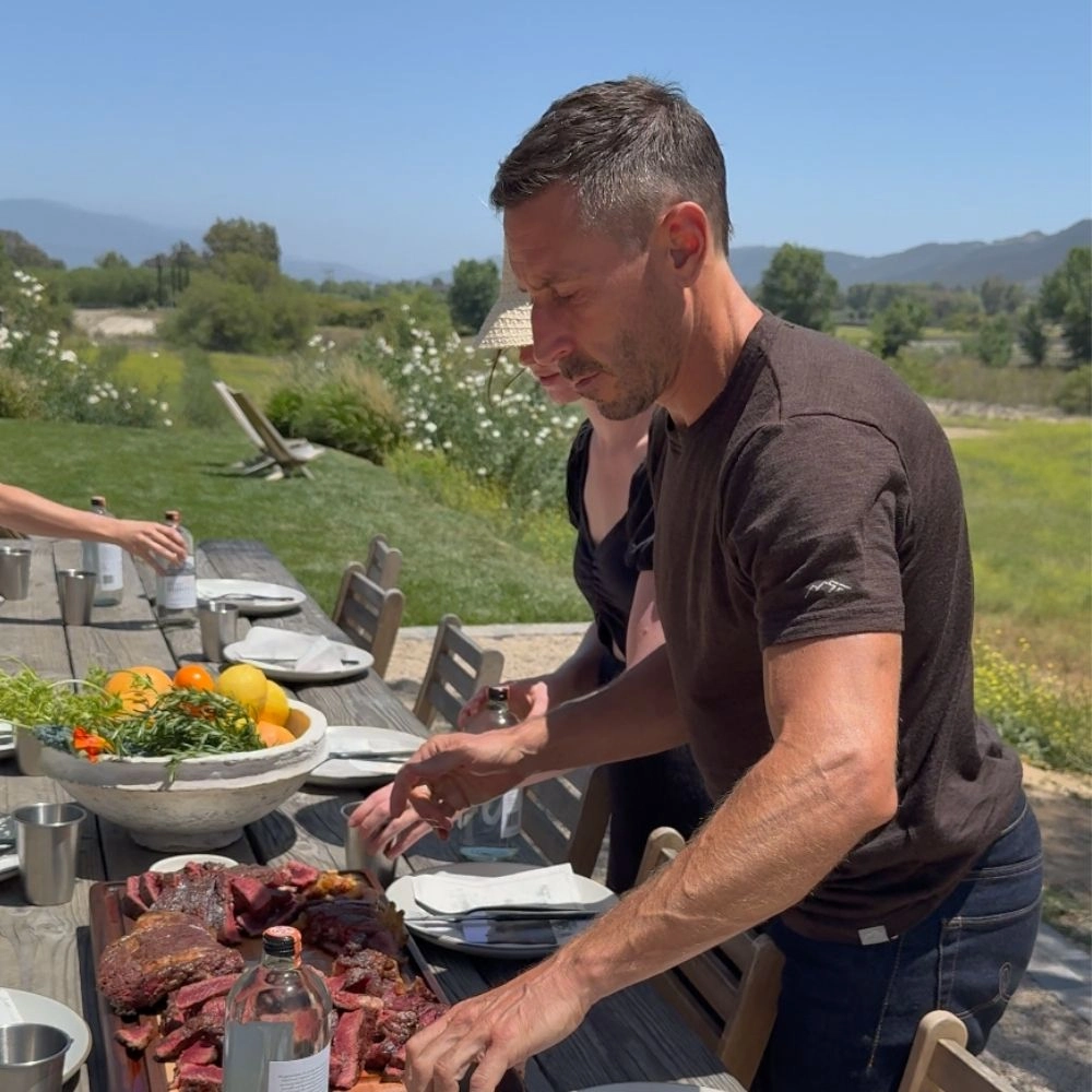 Person arranging slices of meat on a wooden table outdoors.