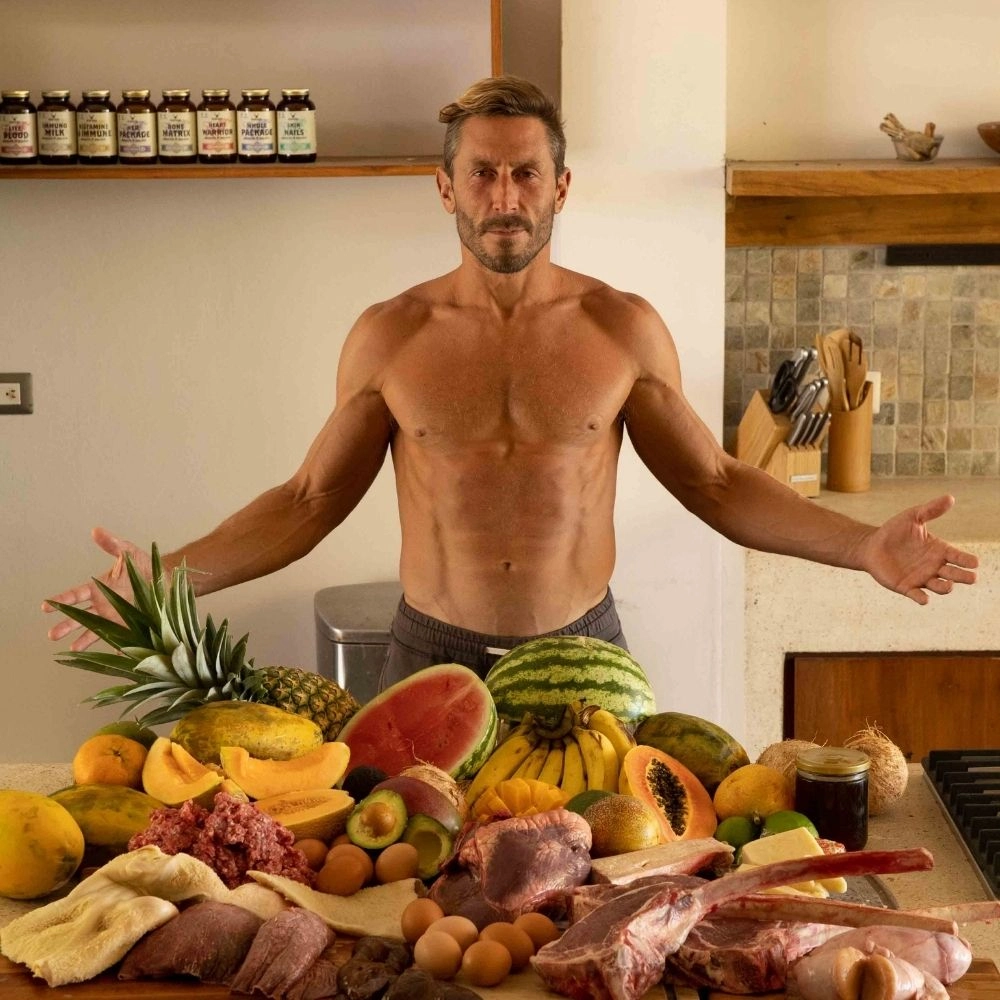 Shirtless man behind a table with fruits, meats, and eggs.