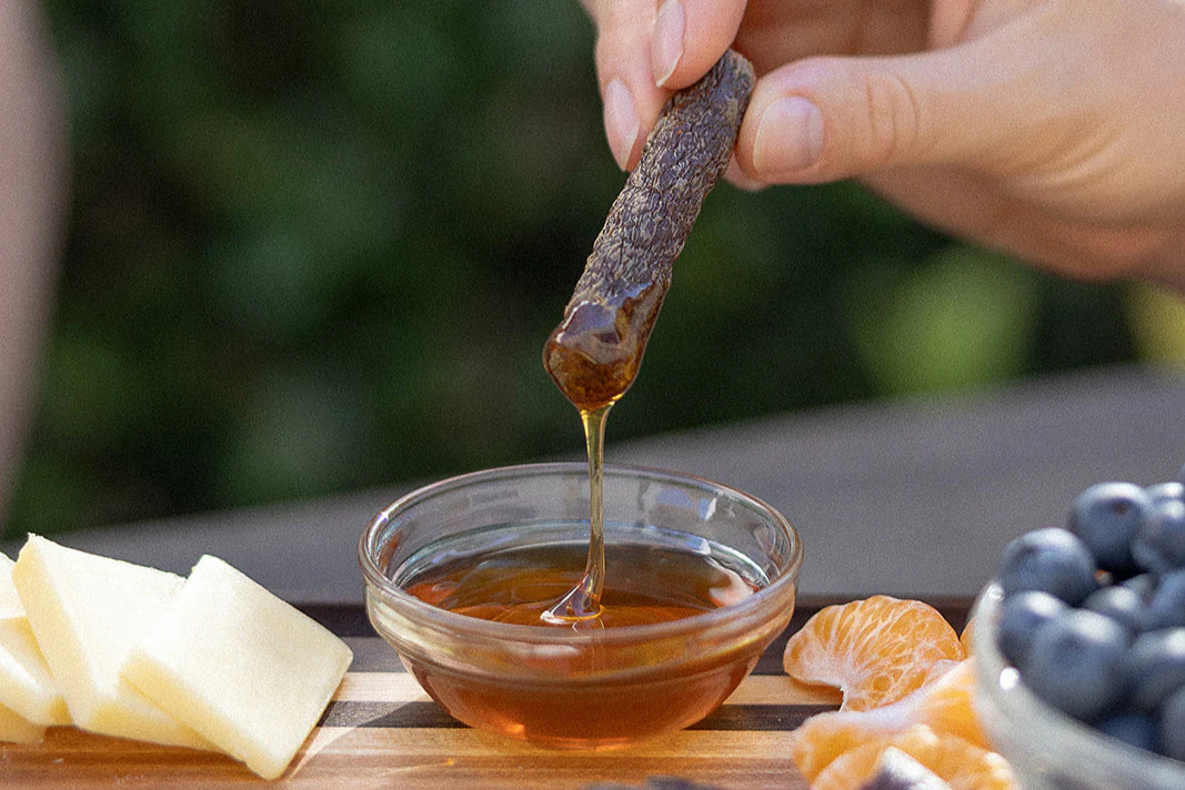 Hand dipping dried meat into honey, with cheese and fruit nearby.