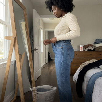A woman with an afro stands in profile, trying on a pair of blue jeans in a bedroom.