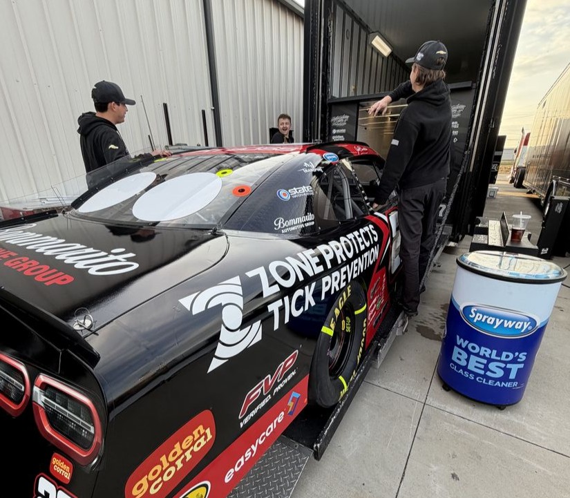 Three crew members work on a black race car, preparing it next to a large transport trailer.