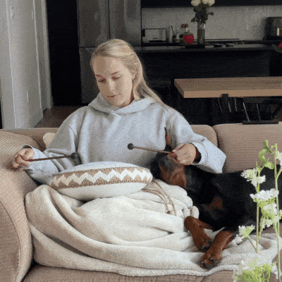 Person on a couch playing a drum with a sleeping dog beside them.
