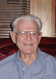 A headshot of a smiling elderly man with white hair and glasses, wearing a blue button-up shirt.