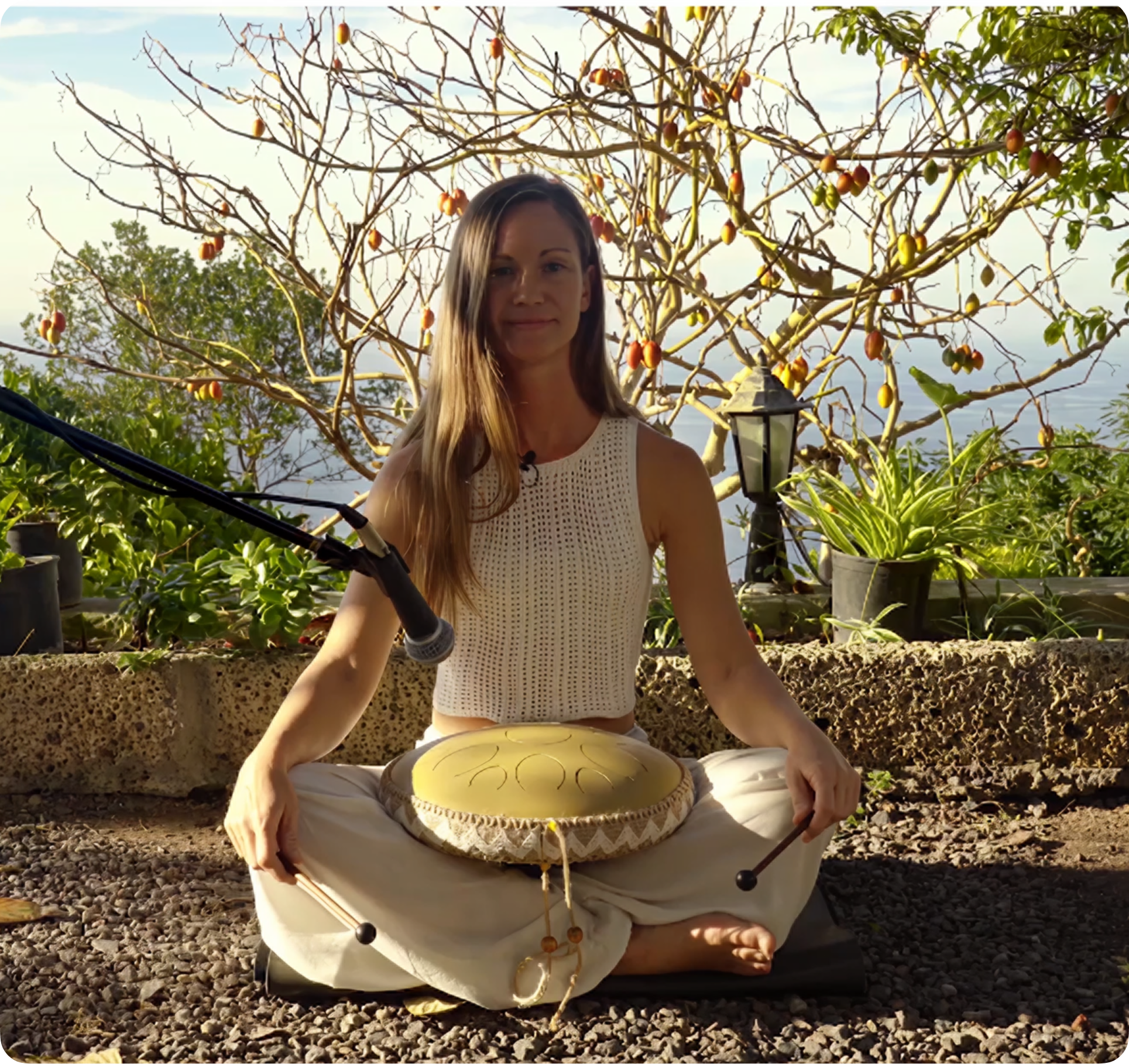 A woman sits cross-legged outdoors with a steel tongue drum in her lap and a microphone in front.