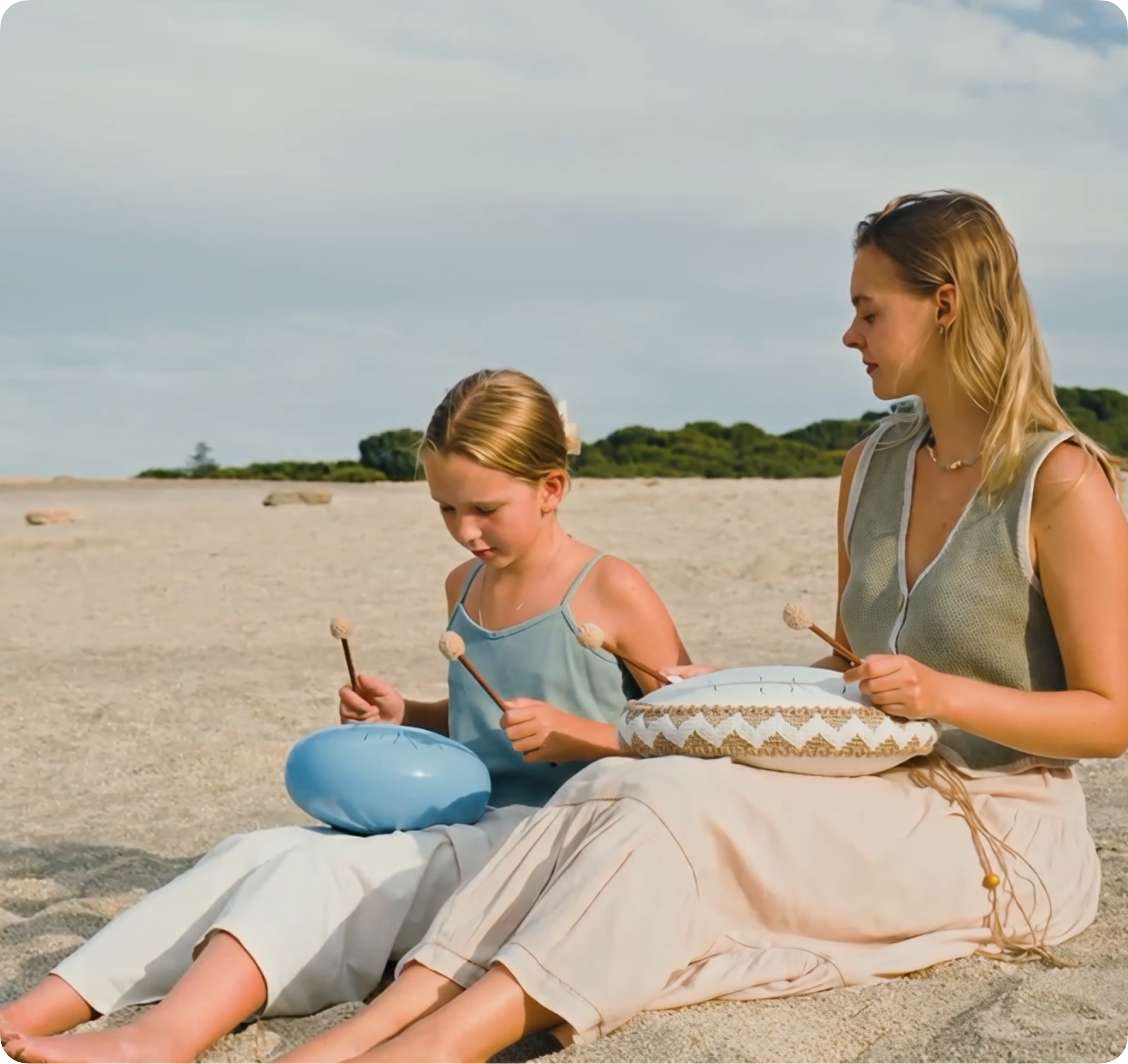 A woman and a girl sit on a sandy beach playing steel tongue drums with mallets.