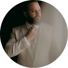 A bearded man in a light-colored suit adjusts his tie while looking to the side, framed in a circle.