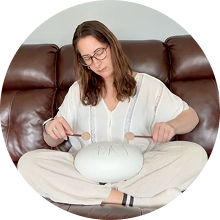 A woman with glasses sits cross-legged on a brown couch playing a white steel tongue drum.