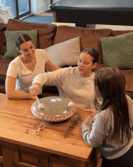 Three people playing a tongue drum on a wooden table in a living room.