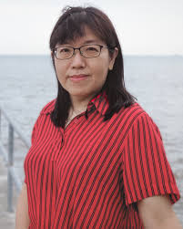 A woman with dark hair and glasses wearing a red and black striped shirt, standing near water.
