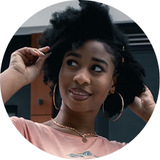 A young Black woman with natural hair smiles and looks to the side while touching a curl.