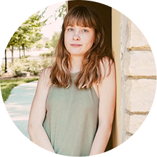 A circular headshot of a young woman with light brown, wavy hair and bangs, wearing a green top outdoors.