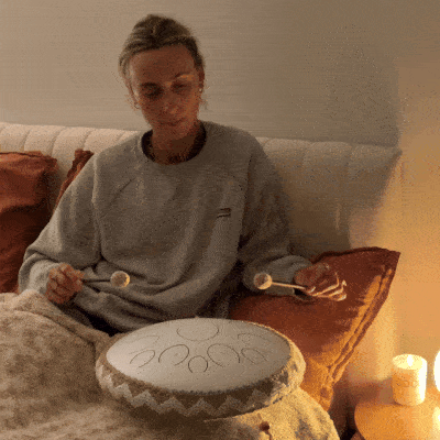 Person playing a handpan drum in a cozy room, with a lit candle on a table.