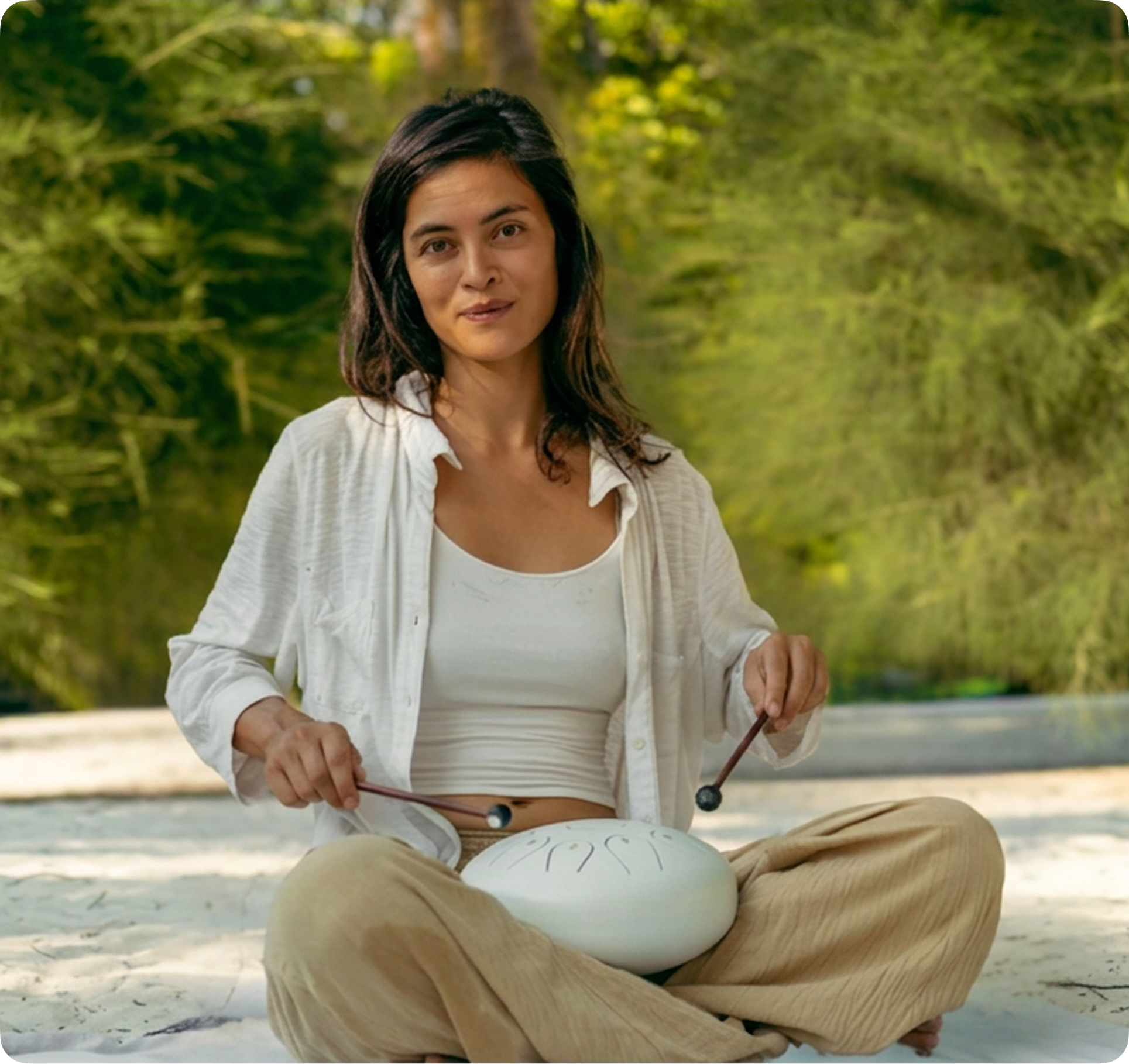 A woman sits cross-legged outdoors, playing a white steel tongue drum with two mallets.