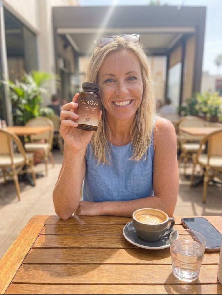 A smiling blonde woman sits at a cafe table holding a bottle of Tanova supplements.
