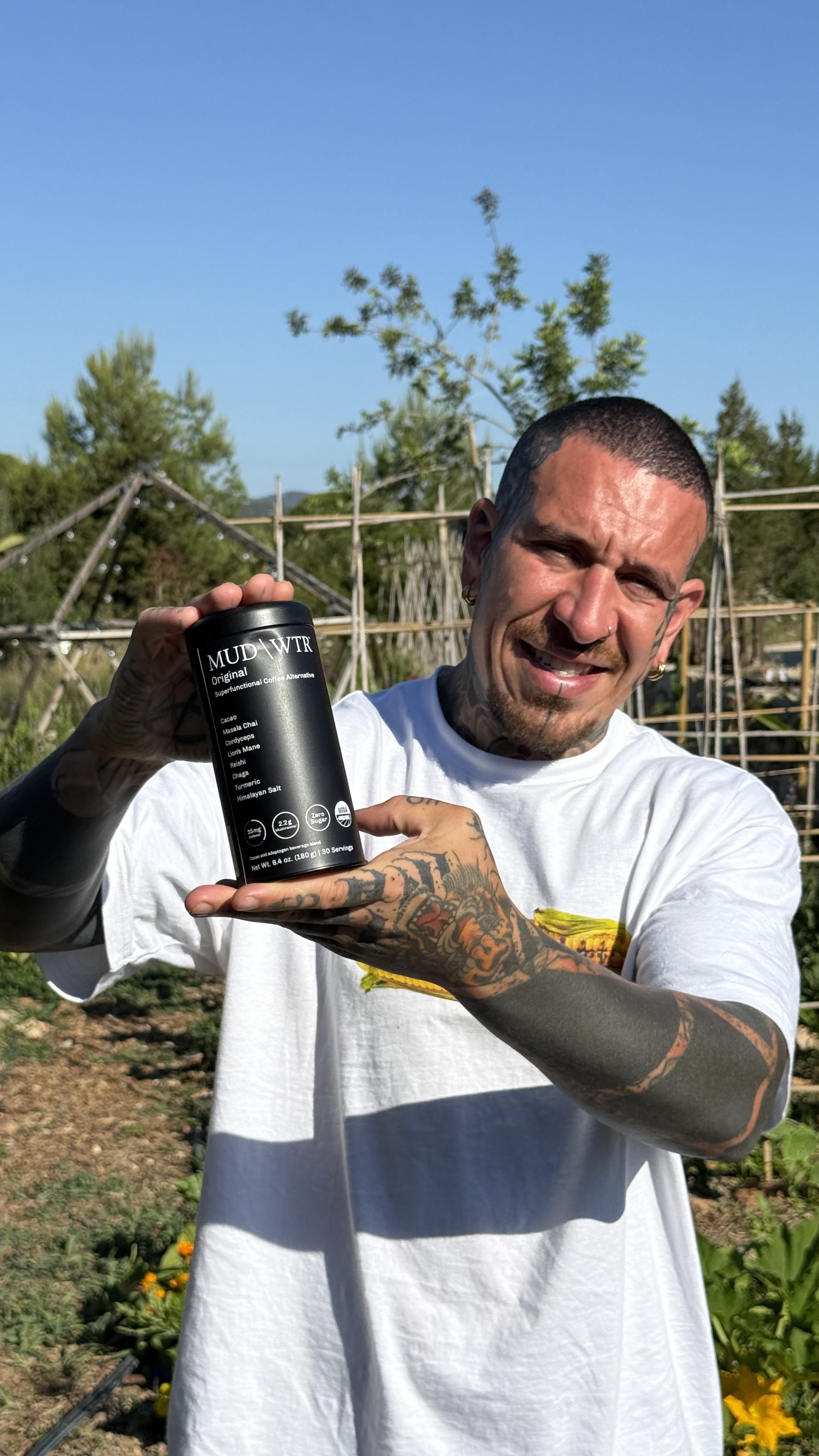 A smiling, tattooed man holds up a black canister of MUD\WTR in a sunny, outdoor garden setting.