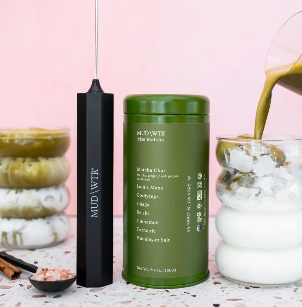 Green canister, black frother, pouring liquid into glass with ice, pink background.