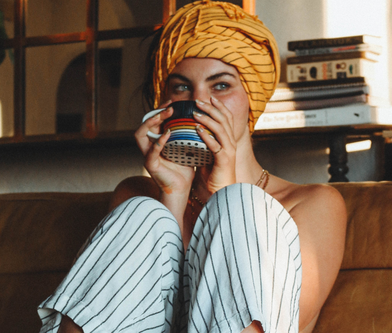 Person with yellow headscarf sipping from a colorful cup, sitting on a couch.