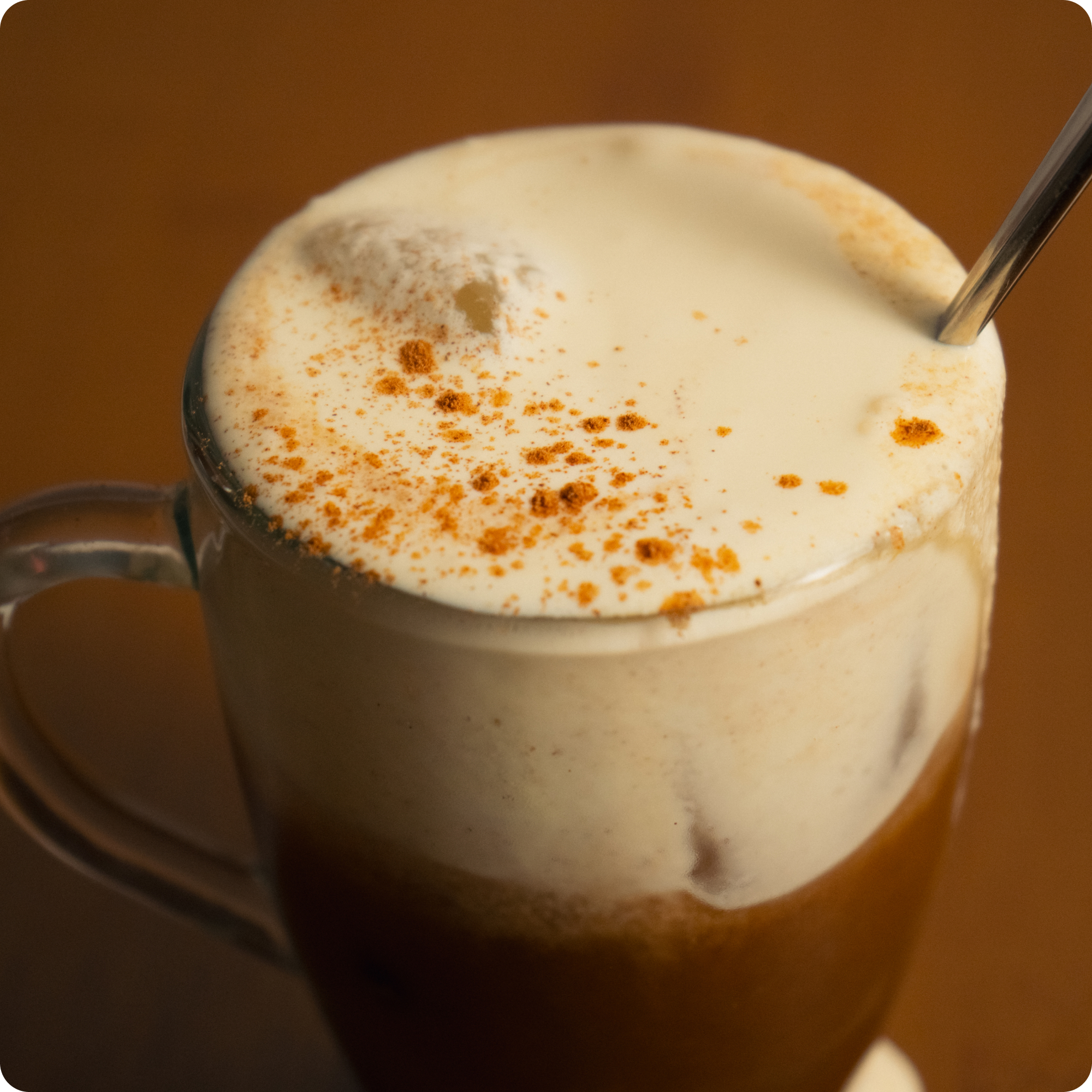 A close-up of a coffee drink in a glass mug, topped with thick foam and spices, with a spoon.