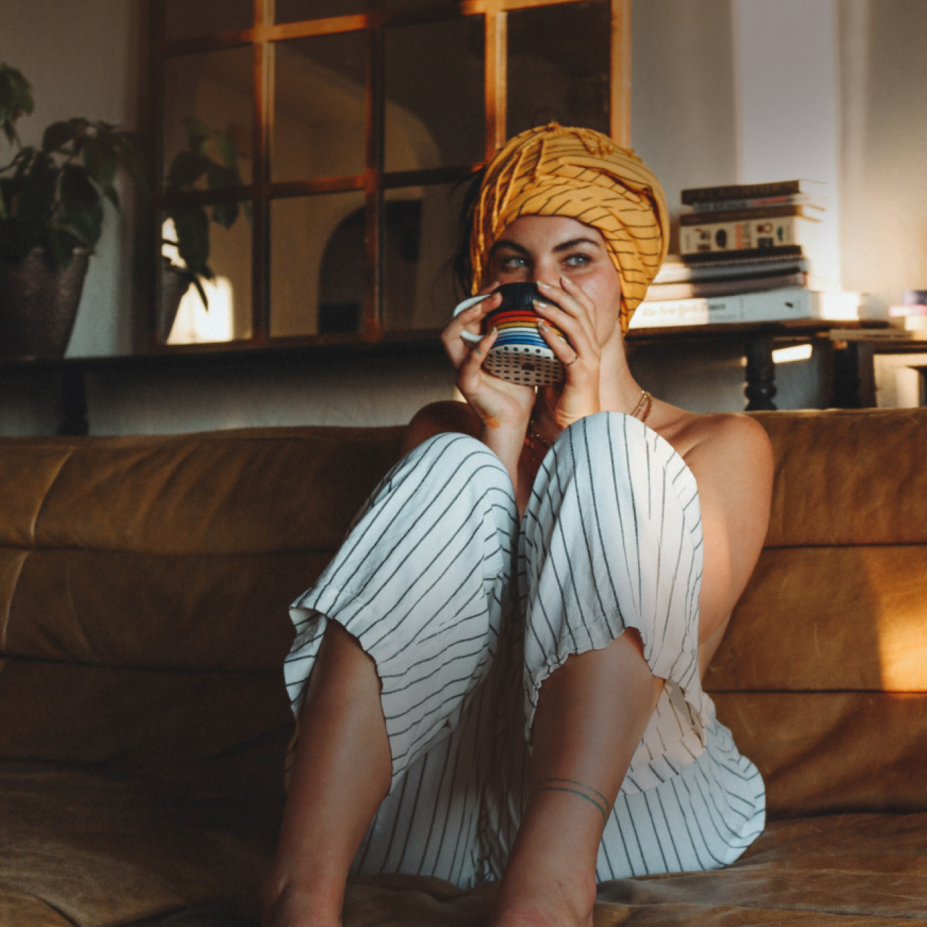 A person in a yellow head wrap sits on a couch, drinking from a colorful mug.