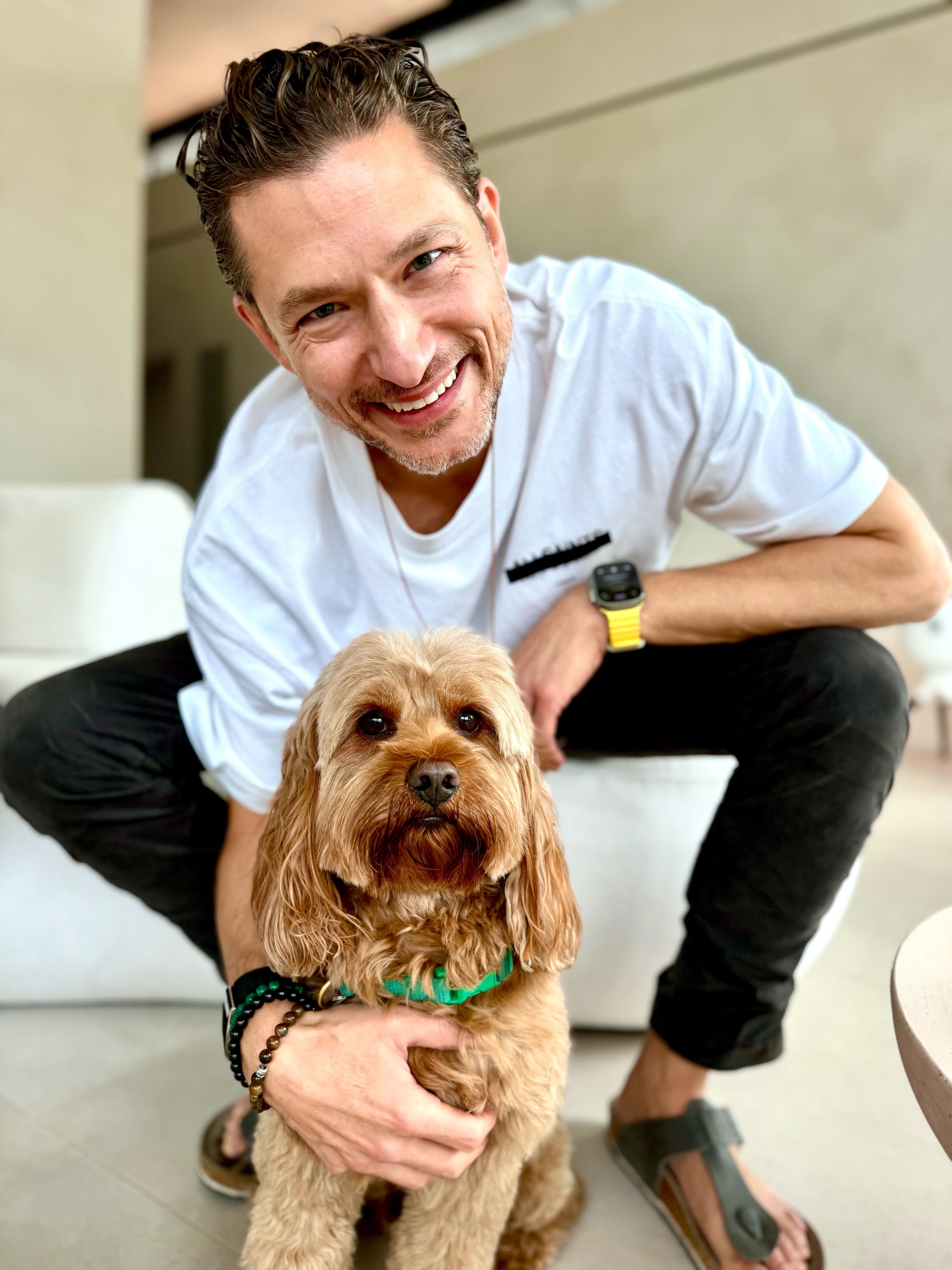 Man smiling while crouching with a small brown dog indoors.