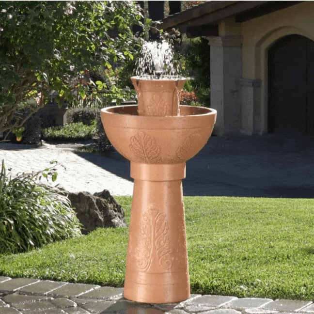 Terracotta fountain with water flowing, set in a garden with grass and stone path.