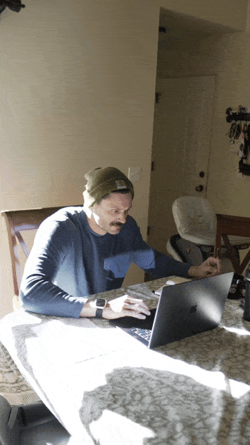 Person in a beanie working on a laptop at a sunlit table.