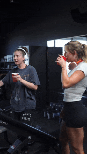 Two women in a gym holding red cups, one talking, the other drinking.