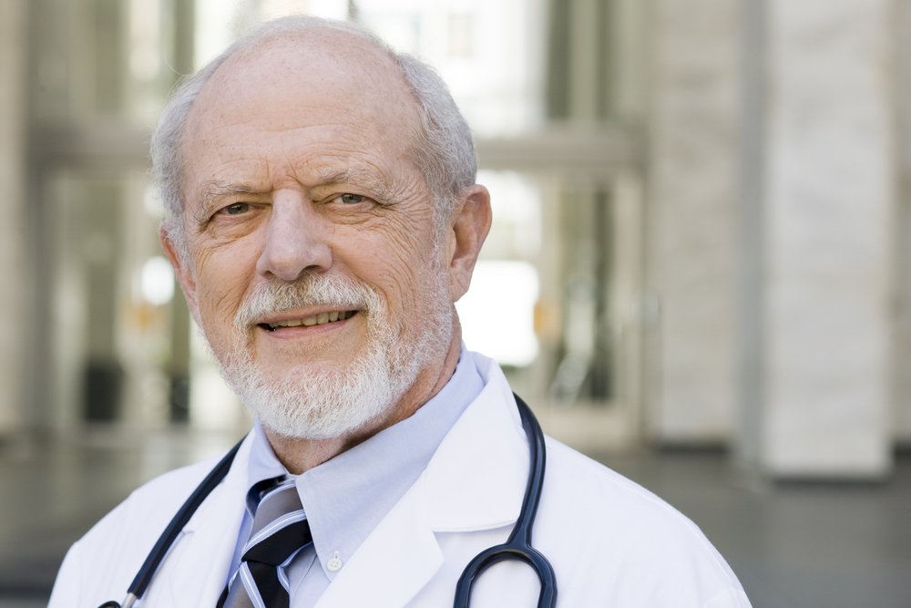 Elderly man in a white coat with a stethoscope, standing outside a building.