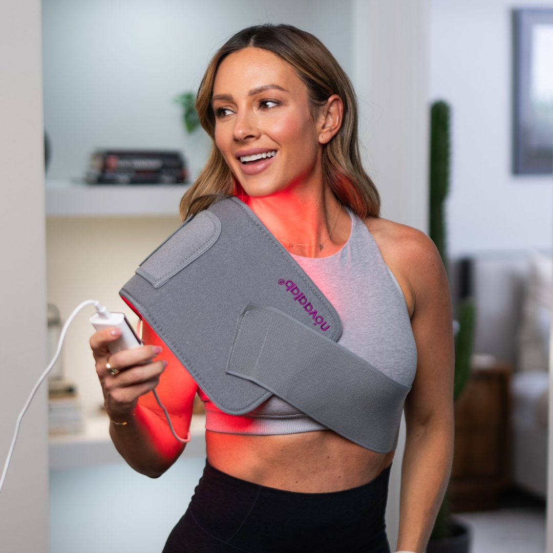 A woman uses a red light therapy wrap on her shoulder while holding the controller.