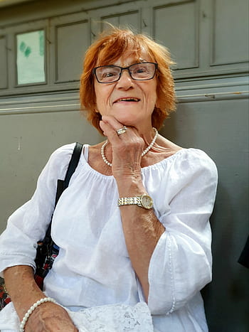 Elderly woman with glasses and white blouse, sitting and smiling.