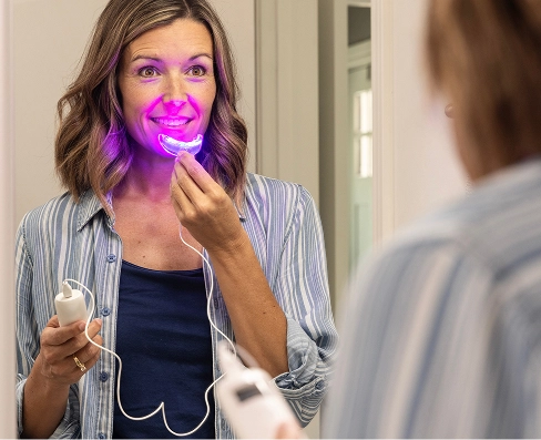A woman looks in a mirror while using a purple light-up teeth whitening device.