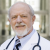Elderly man with a beard wearing a white coat and stethoscope, standing outdoors.