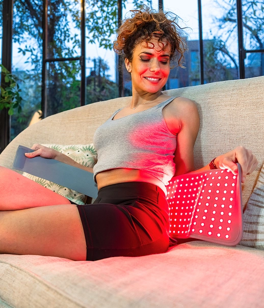 A woman sits on a couch, holding a glowing red light therapy panel against her lower back.