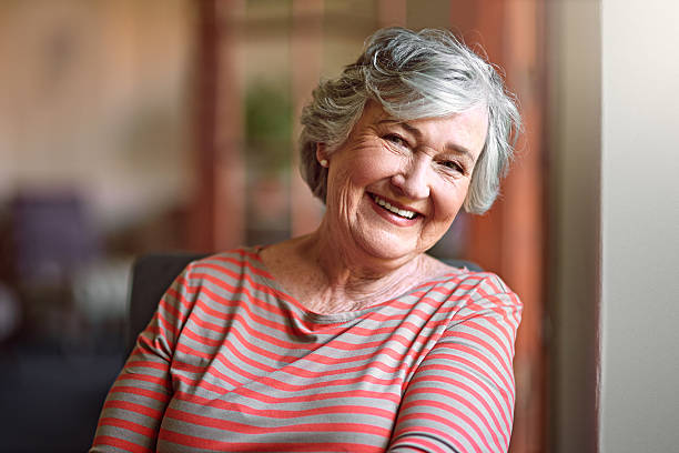 Smiling elderly woman with gray hair in a striped shirt, sitting indoors.