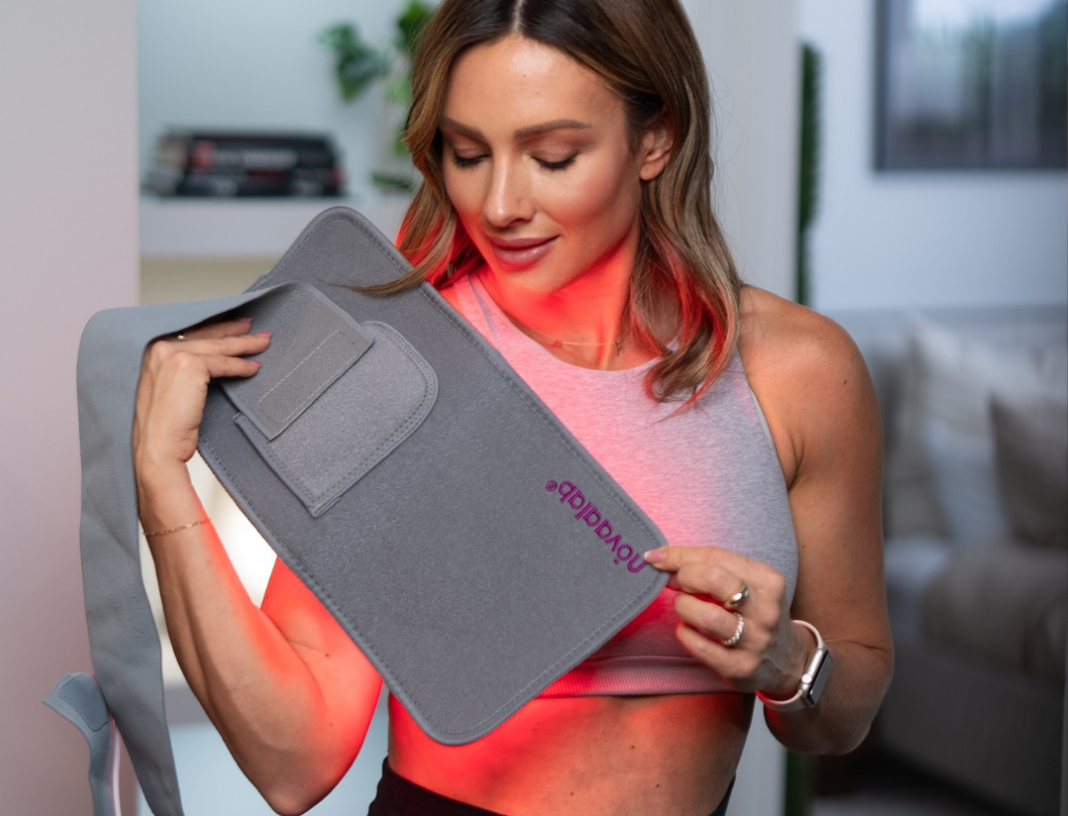 A woman holds a grey, flexible red light therapy device that is glowing red against her body.