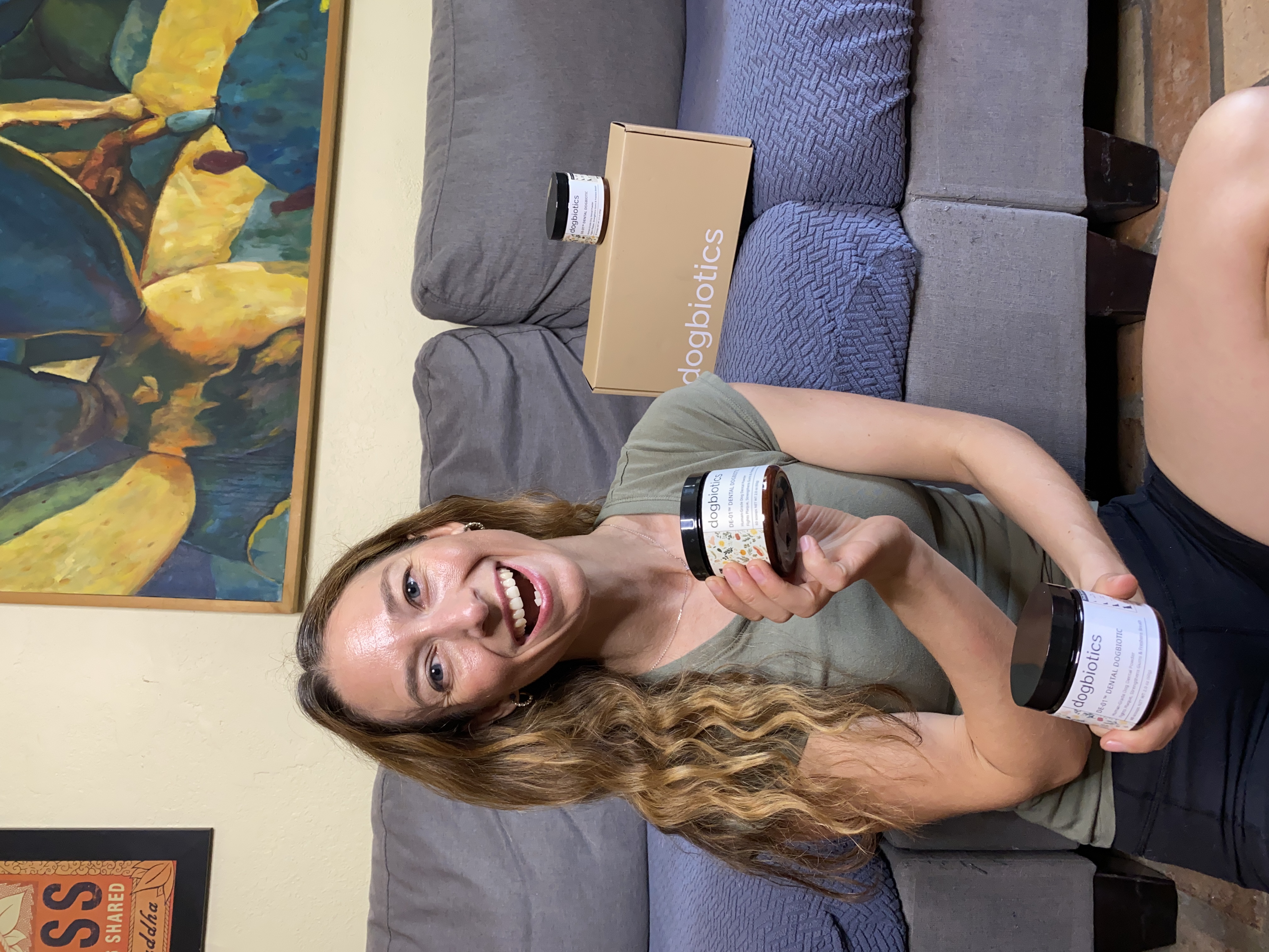 Smiling person holding products labeled 'dogbiotics' near a couch with colorful artwork behind.