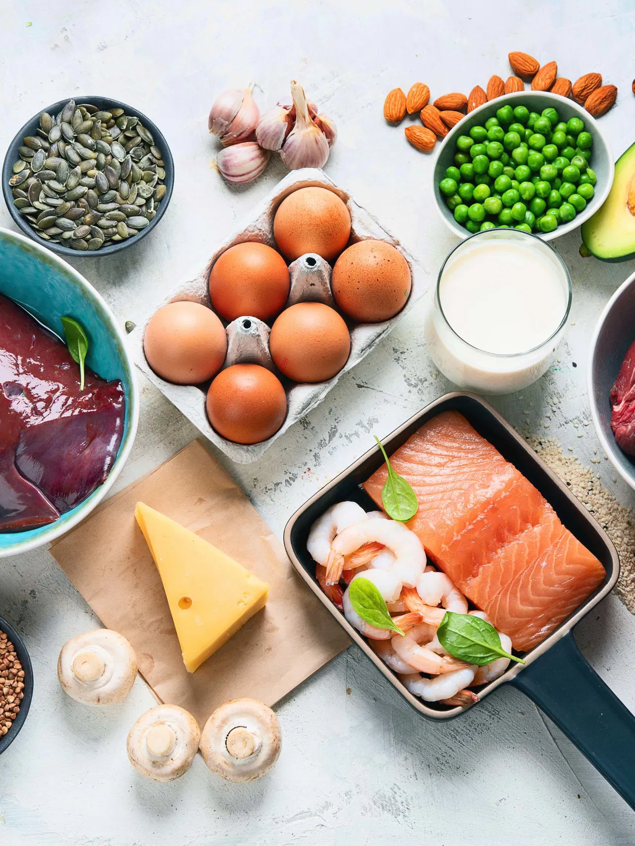 Variety of foods including eggs, salmon, shrimp, cheese, mushrooms, avocado, and nuts on a light surface.